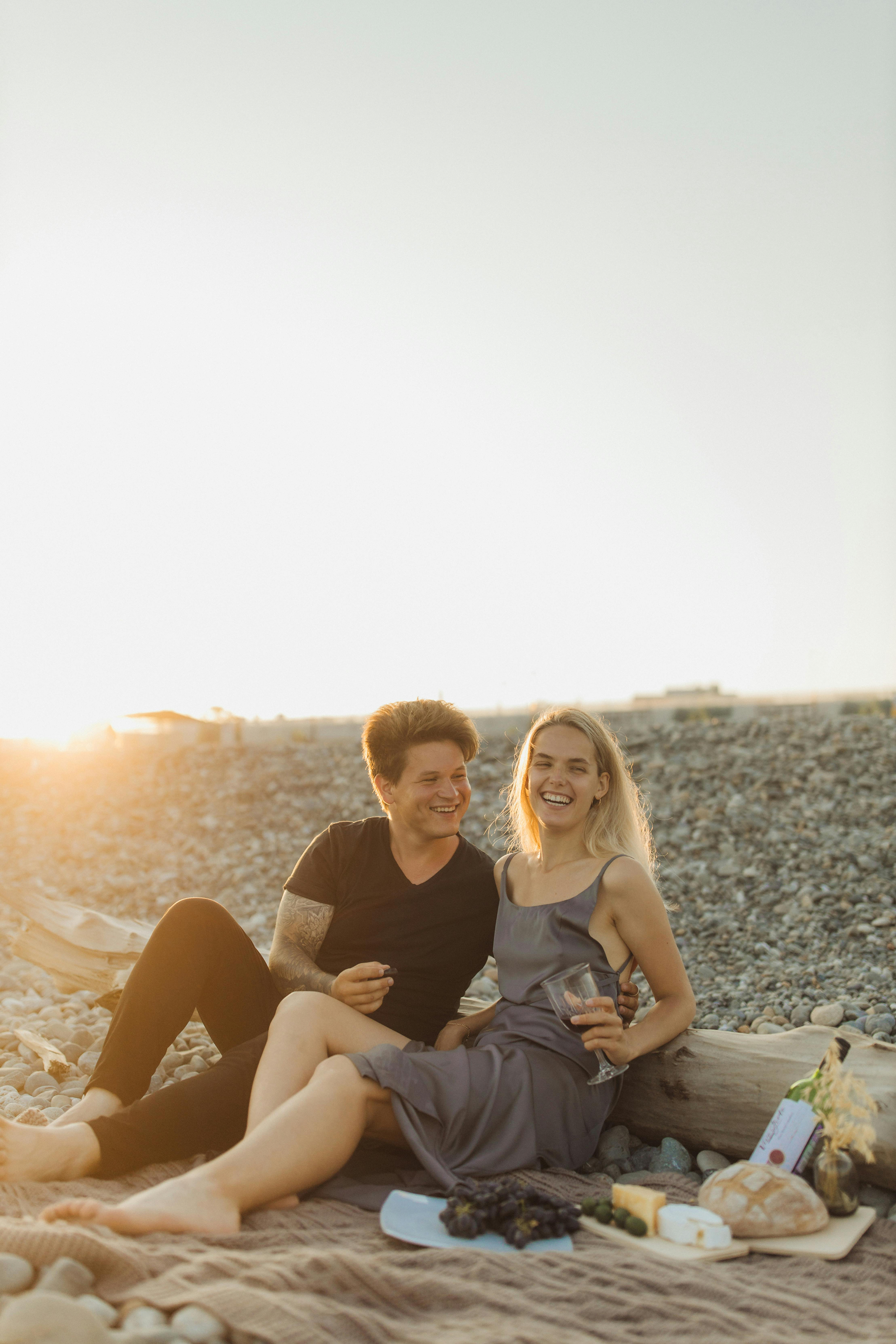 Ett leende par sitter tillsammans på en stenig strand i solnedgången, en håller ett glas.