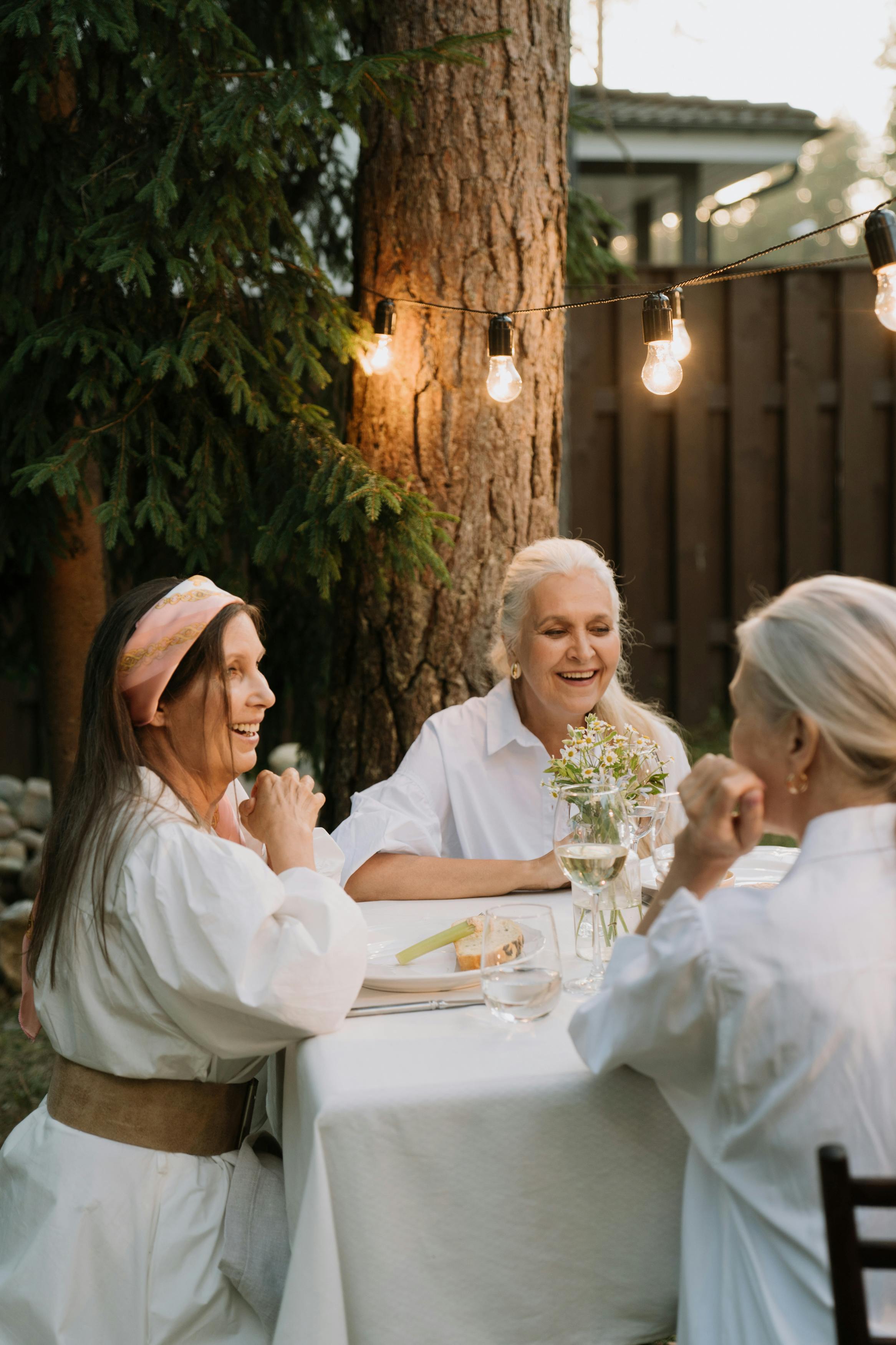 Tre kvinnor skrattar tillsammans vid ett utomhusbord med blommor i en vas.